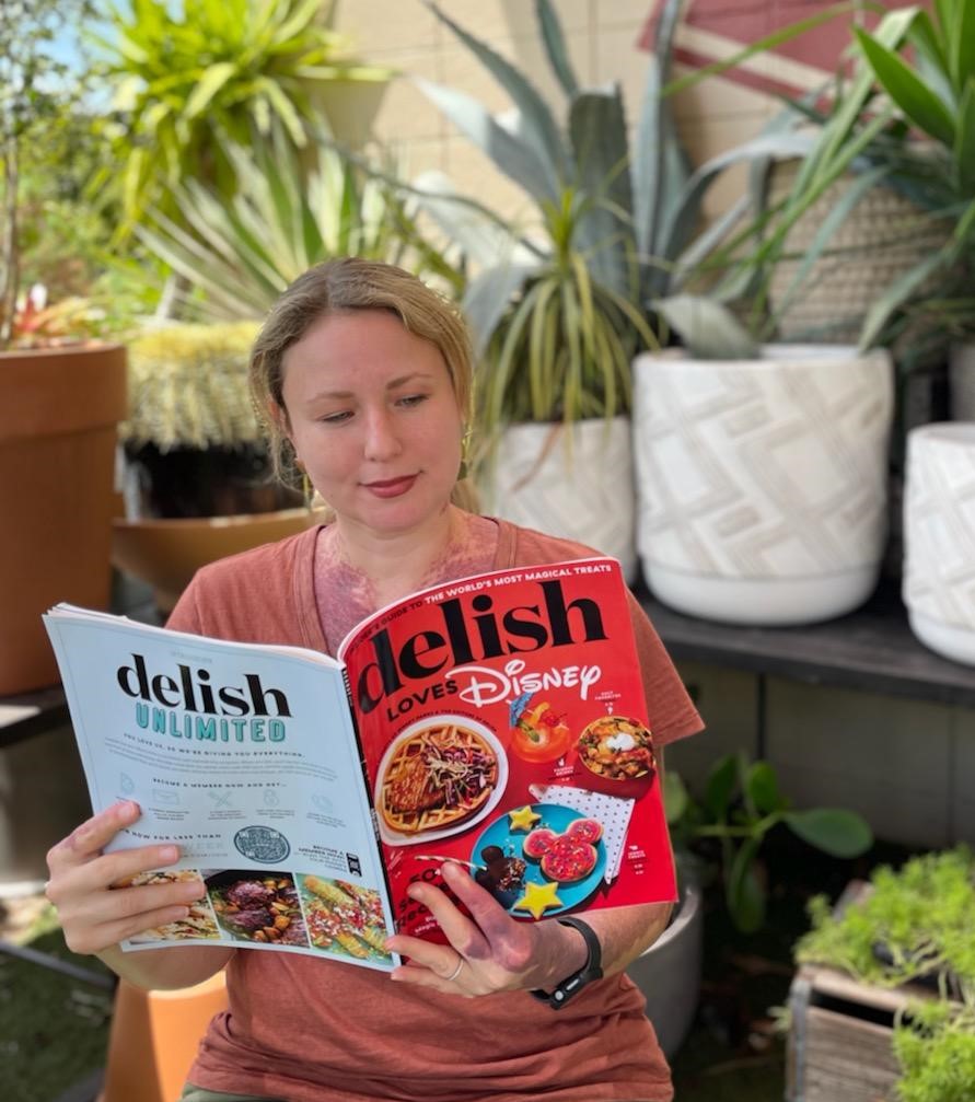 girl with blonde hair with orange shirt holding a red cover magazine in front of green plants