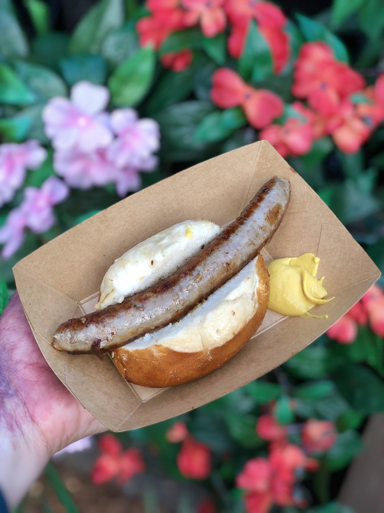 Brown sausage in a bun on a brown plate in front of pink flowers.