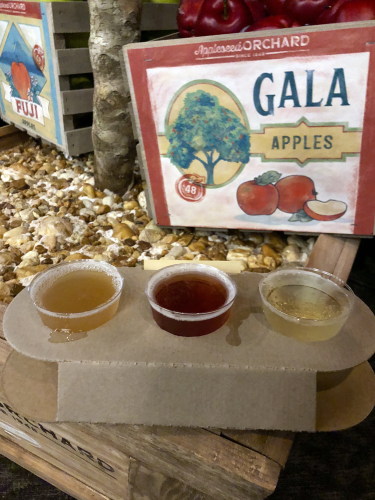 Three yellow ciders in plastic cups in front of a red sign.