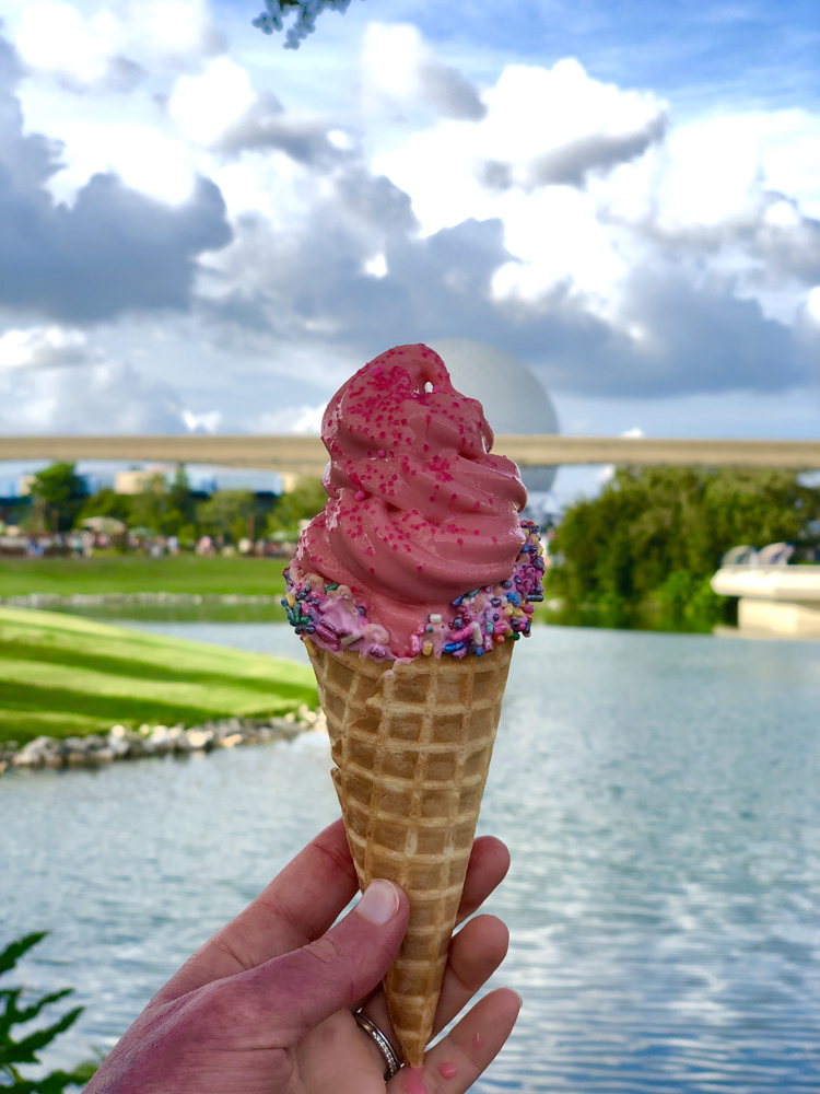 Pink ice cream in a cone in front of water and blue sky.