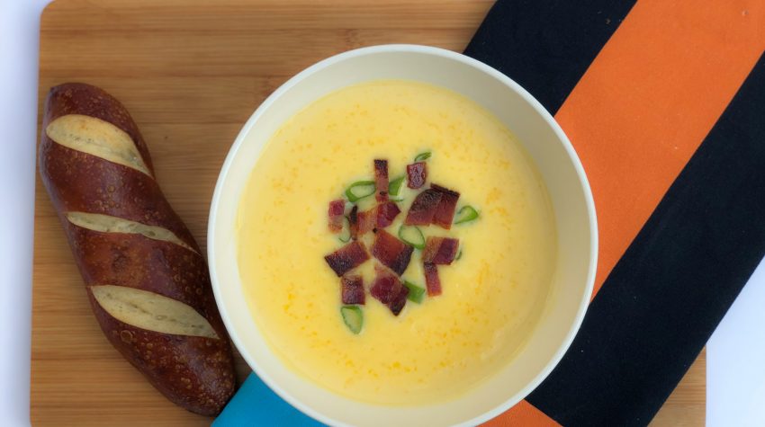 White bowl of cheese soup on a brown board with brown bread and orange and blue kitchen towel