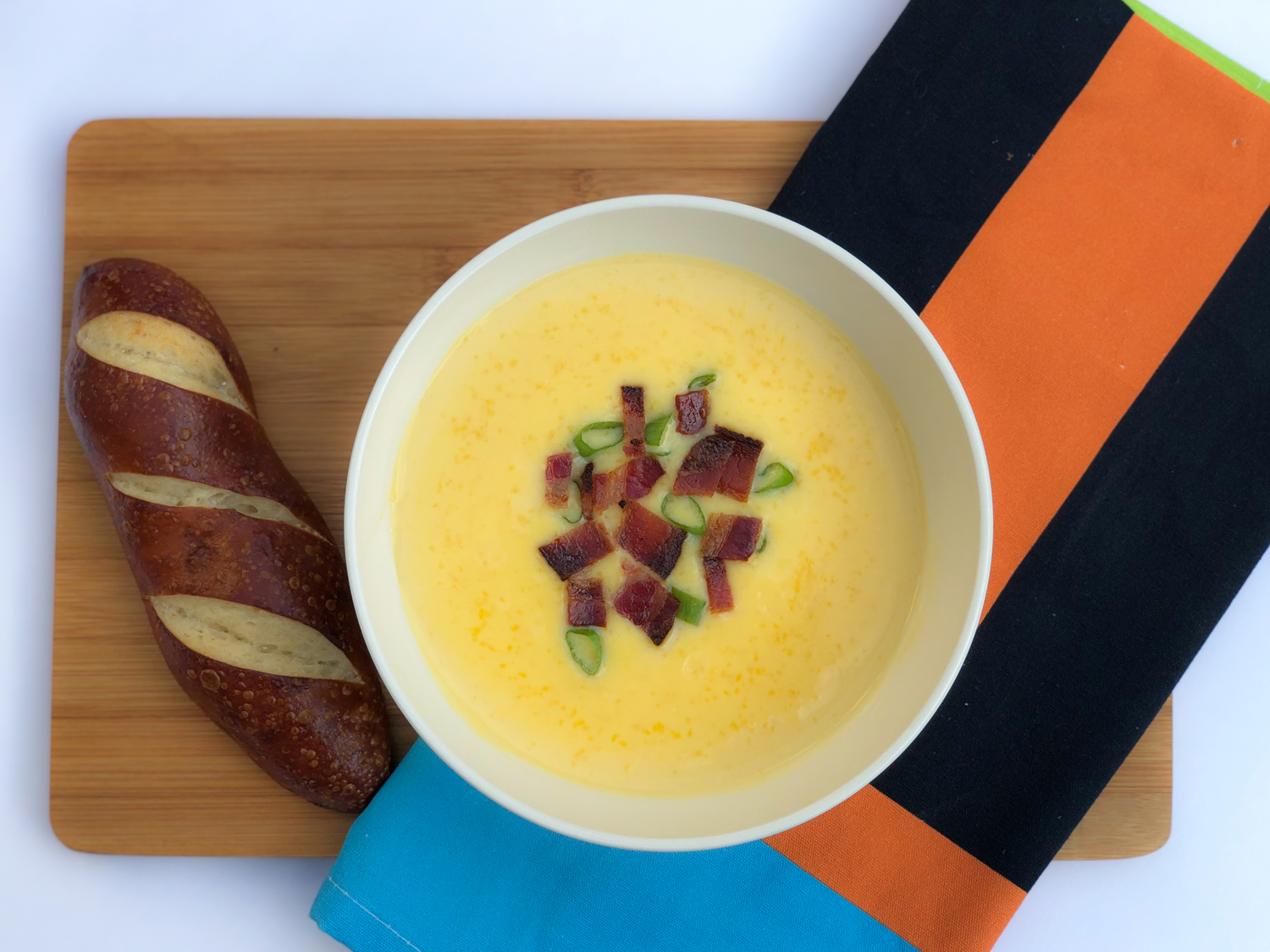 White bowl of cheese soup on a brown board with brown bread and orange and blue kitchen towel