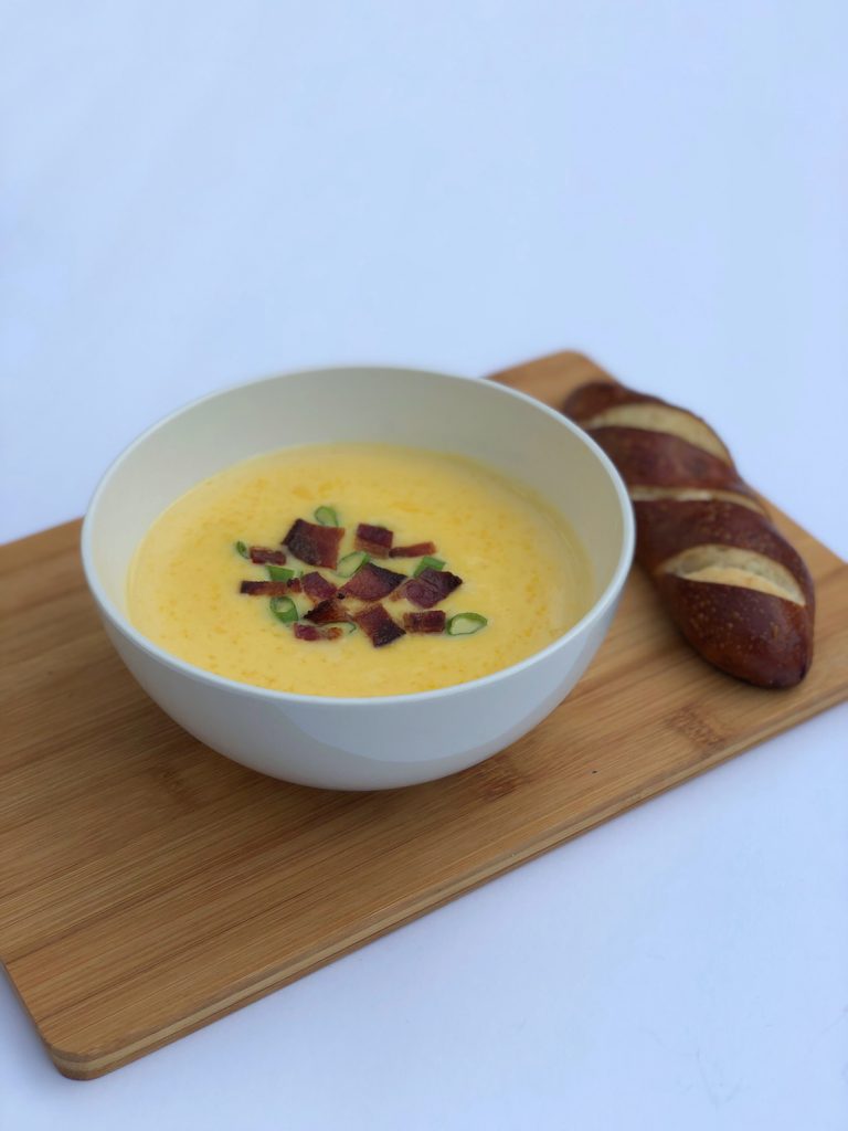 White bowl of cheddar cheese soup on a brown board with brown bread on a white background