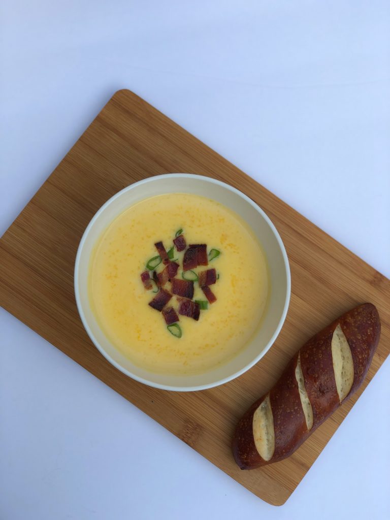 White bowl of cheddar cheese soup on a brown board with brown bread on a white background