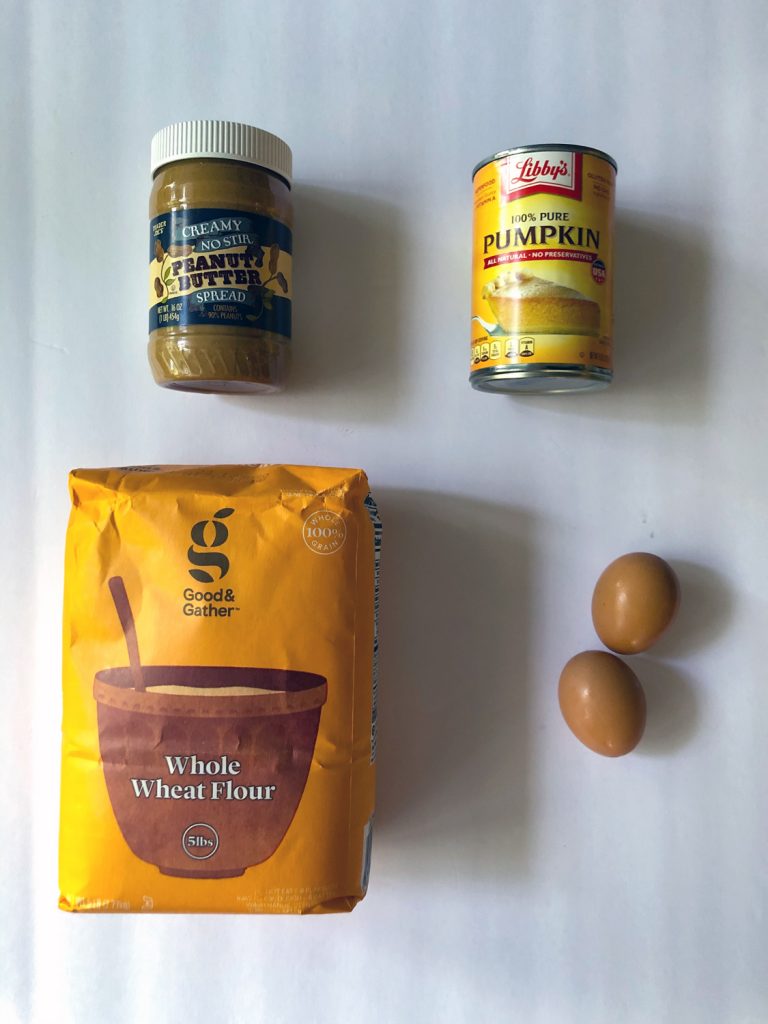 Jar of peanut butter, can of pumpkin puree, orange bag of whole wheat flour and two brown eggs on a white background.